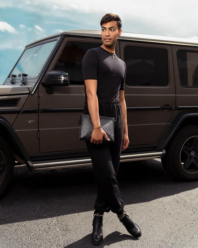 Pollito Tropical in a black t-shirt and pants in front of a black luxury vehicle.